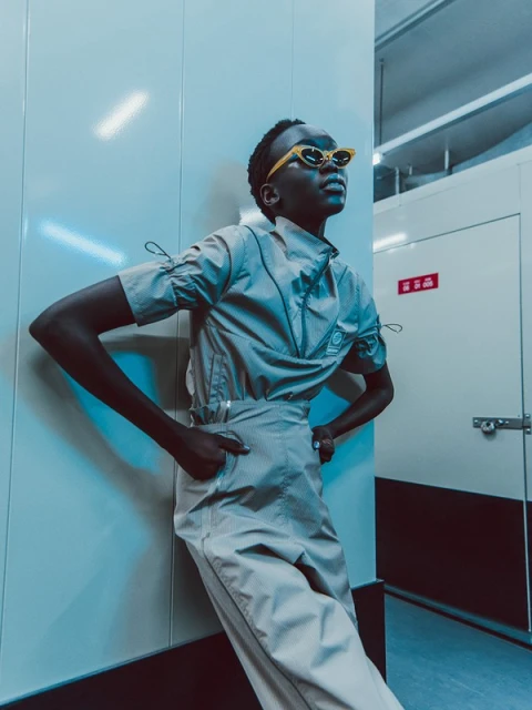 Stylish woman in beige two-piece outfit and yellow sunglasses poses against an indoor white wall with a modern, fashionable look.