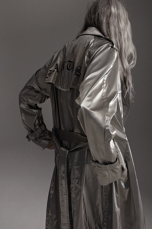 Back view of an adult wearing a silver trench coat with black text design, standing indoors against a gray background in a studio shot.
