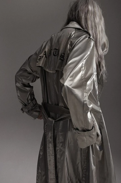 Back view of an adult wearing a silver trench coat with black text design, standing indoors against a gray background in a studio shot.