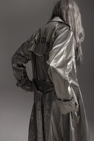 Back view of an adult wearing a silver trench coat with black text design, standing indoors against a gray background in a studio shot.