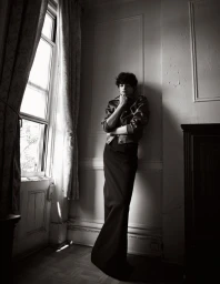 Monochrome portrait of a person leaning against a wall by a window wearing a leather jacket and long skirt