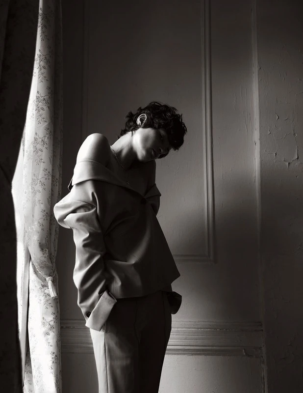 Model with short dark hair wearing an off-the-shoulder top standing indoors near a curtained window.