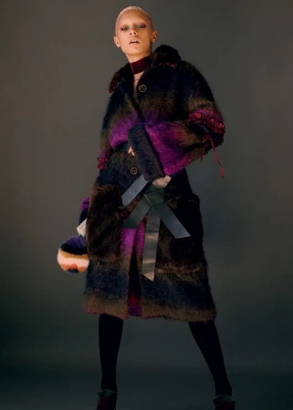 Model with short buzzed hair wearing a plush faux fur coat in purple and black tones against a neutral dark grey background.