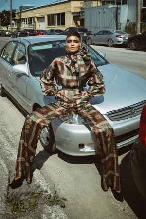 Model wearing patterned silk headscarf and matching outfit perched on the hood of a parked silver car on an urban street.