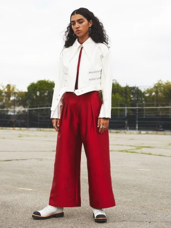 Model standing outdoors wearing a white cropped jacket and shiny red pleated trousers with white boots