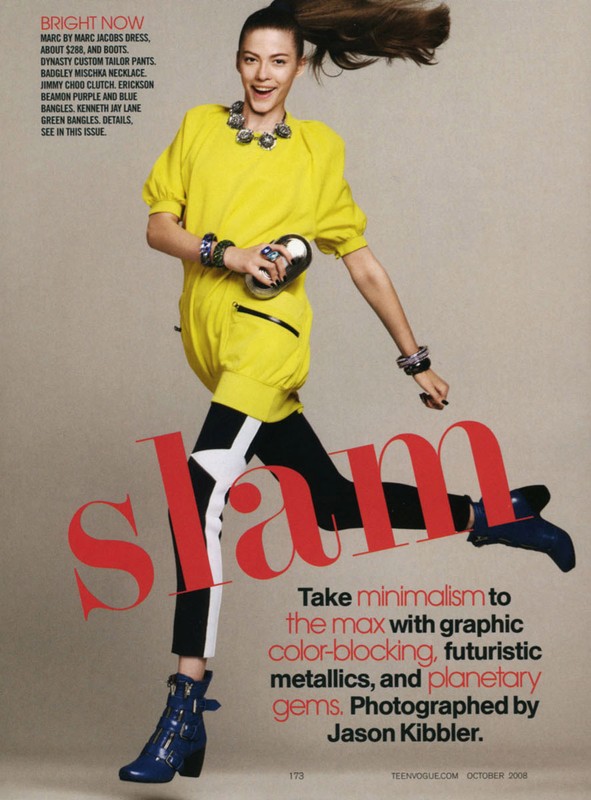 Woman in bright yellow mini dress, black and white pants, and blue booties strikes a pose with a playful expression, indoors.