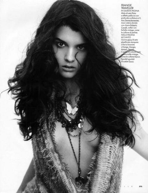 Black and white studio portrait of a female fashion model with wavy hair wearing an open weave top and multiple necklaces.