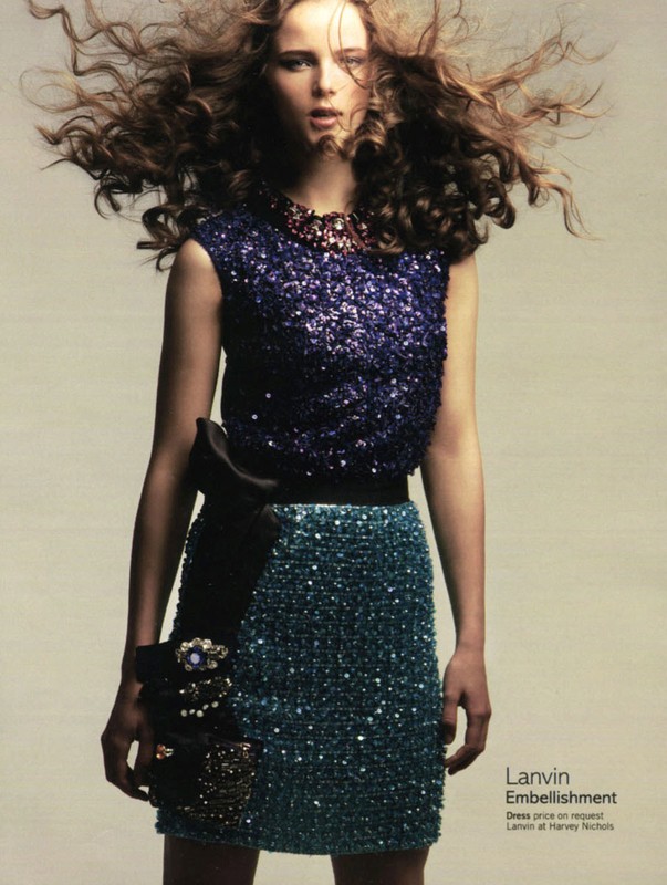 Fashion model in a sequin top and skirt posing in a studio setting with wind-blown curly brown hair for a clothing advertisement.
