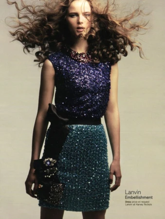 Fashion model in a sequin top and skirt posing in a studio setting with wind-blown curly brown hair for a clothing advertisement.