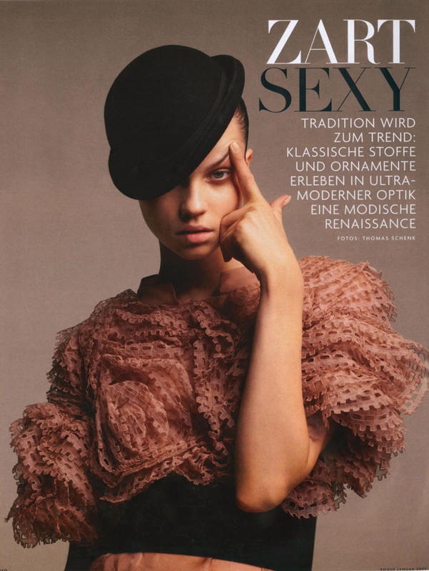 Fashion shot of a young adult woman with dark hair wearing a black hat and frilled top, striking a pose with hand near her forehead.