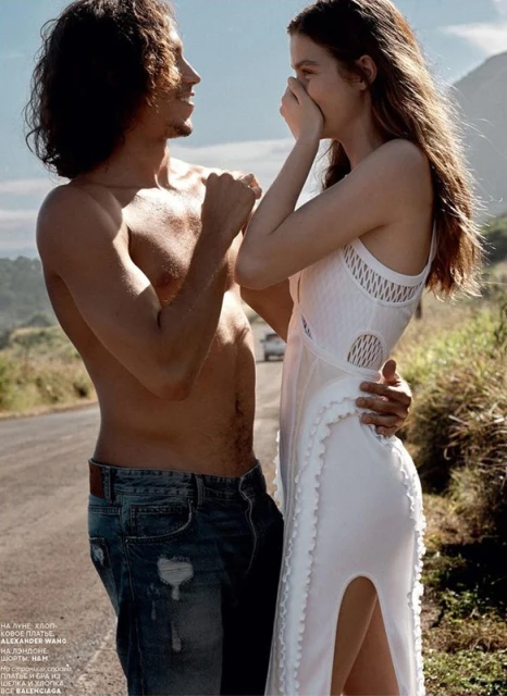 Editorial shot of a smiling couple on a road. The man, shirtless in jeans, has his arm around a woman in a long, white crochet dress.