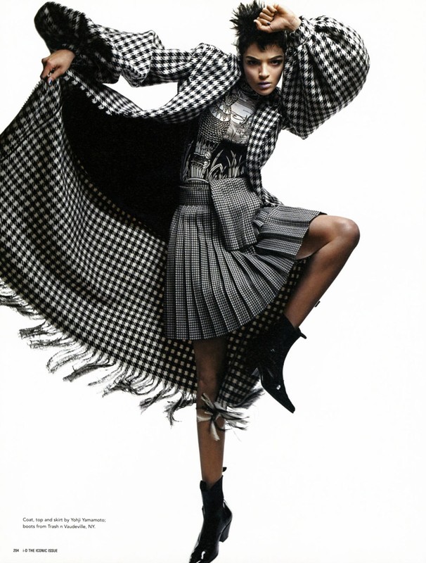 Fashion editorial shot of a woman in a black and white checkered coat and skirt, high-heeled boots, and hand raised against a white backdrop.