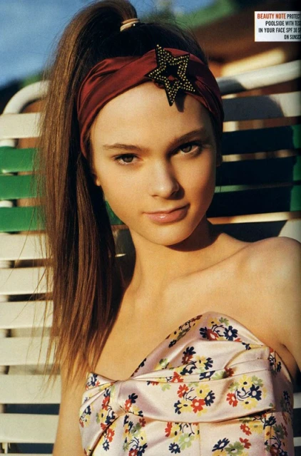 Portrait of a young woman with a floral print top and headband with a star, posing in an outdoor setting with slatted furniture.