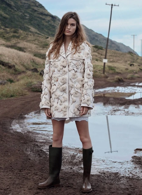 Fashion shot of a woman in a floral coat and boots standing in a muddy field with mountains in the background, creating a unique outdoor scene.