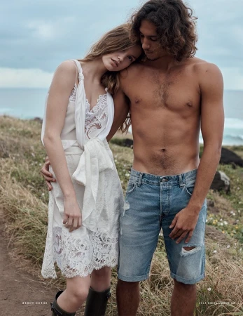A couple standing outdoors near a coastal grassy cliff, with the woman resting her head on the man's shoulder.