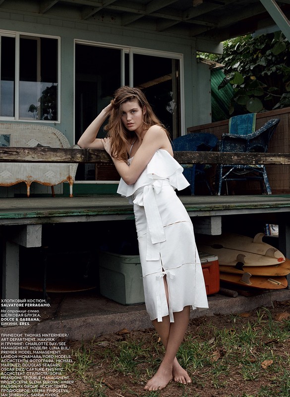 A stylish woman in a white dress poses on a porch with surfboards. Soft, natural light highlights the outdoor fashion shot.