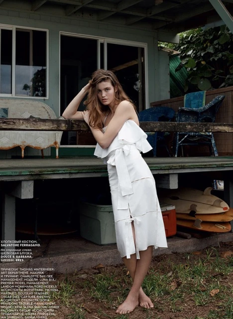A stylish woman in a white dress poses on a porch with surfboards. Soft, natural light highlights the outdoor fashion shot.
