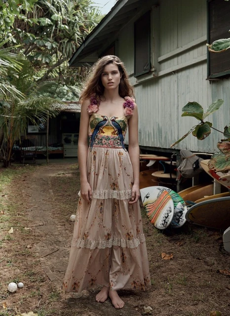 A woman with long brown hair poses in a long floral dress, standing barefoot outside near a rustic building and trees in natural light.