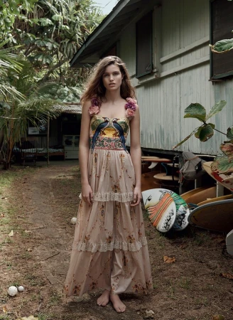A woman with long brown hair poses in a long floral dress, standing barefoot outside near a rustic building and trees in natural light.