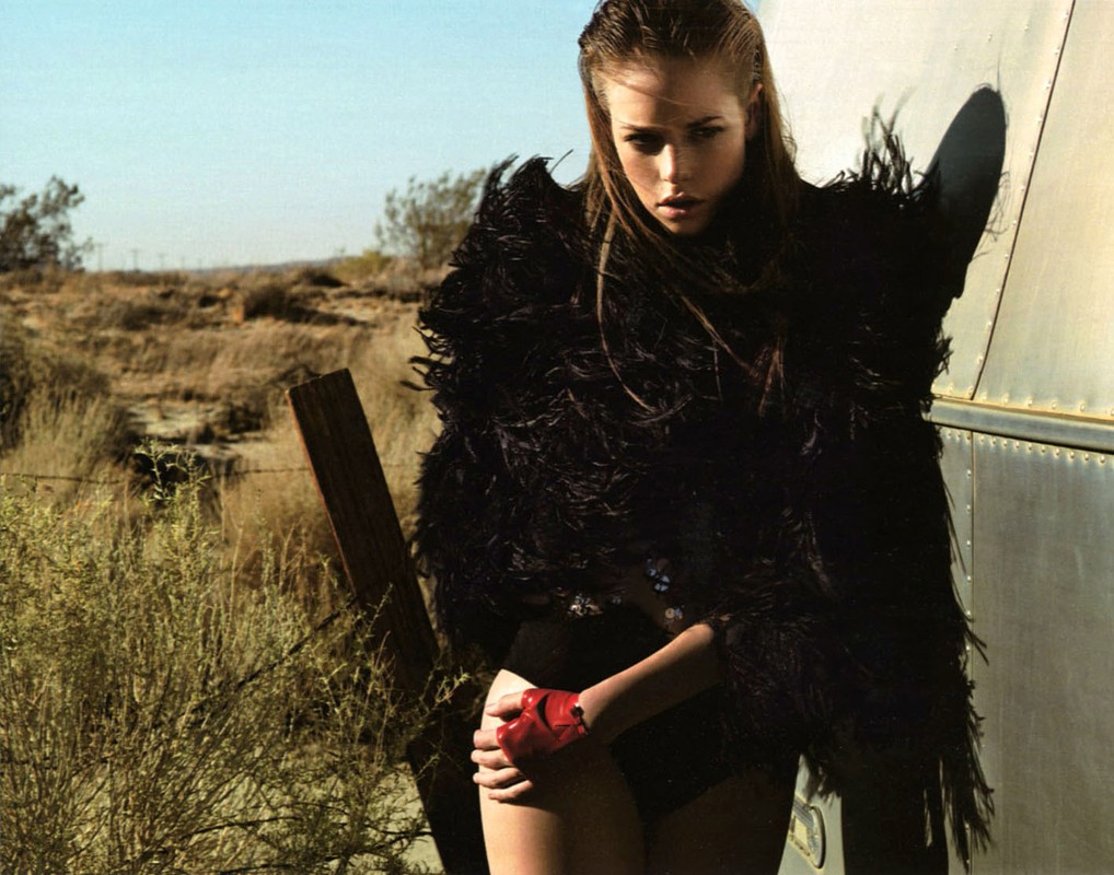 Blonde woman in a feathered jacket poses outside next to a silver trailer. She has red gloves on and wears minimal makeup.