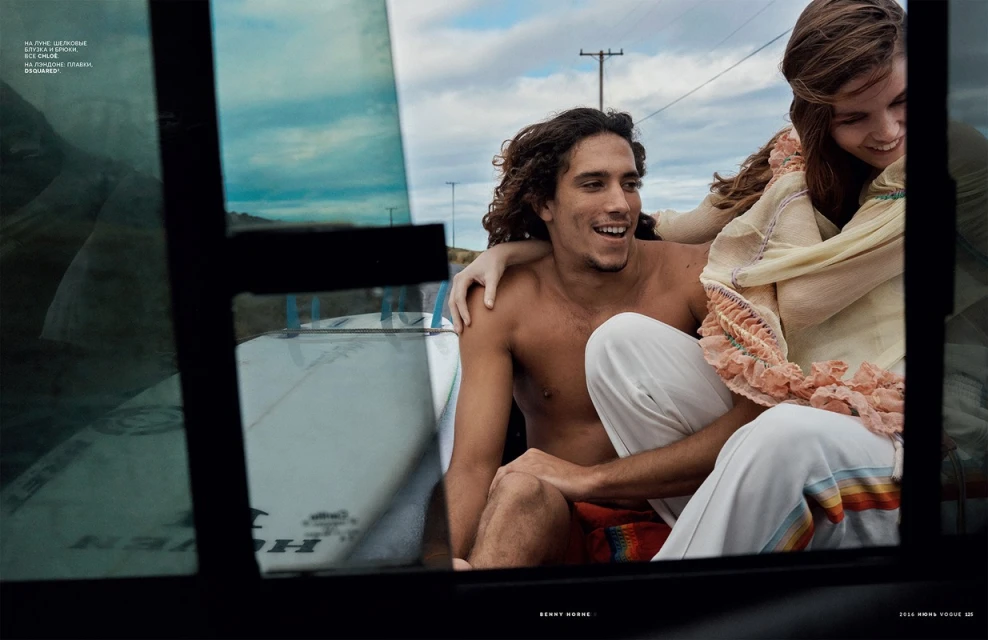 A curly-haired man and a young woman are riding in the back of a pickup truck with surfboards, with a clear sky and power lines visible.