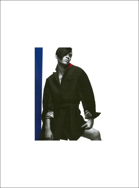 Fashion photo of a person in a belted black coat, posing in a white studio with a vertical blue stripe on the left.