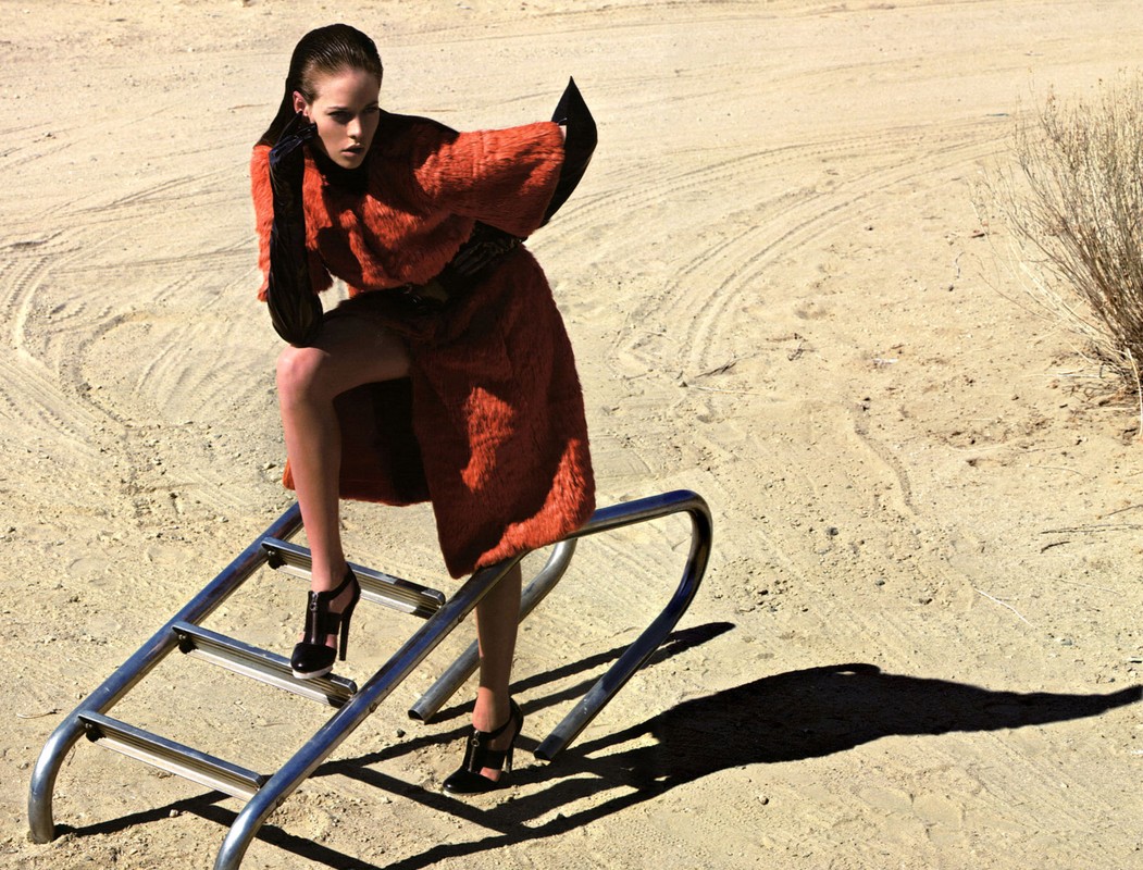 A woman in an orange coat and black heels poses on playground equipment in a desert environment with a neutral color palette.