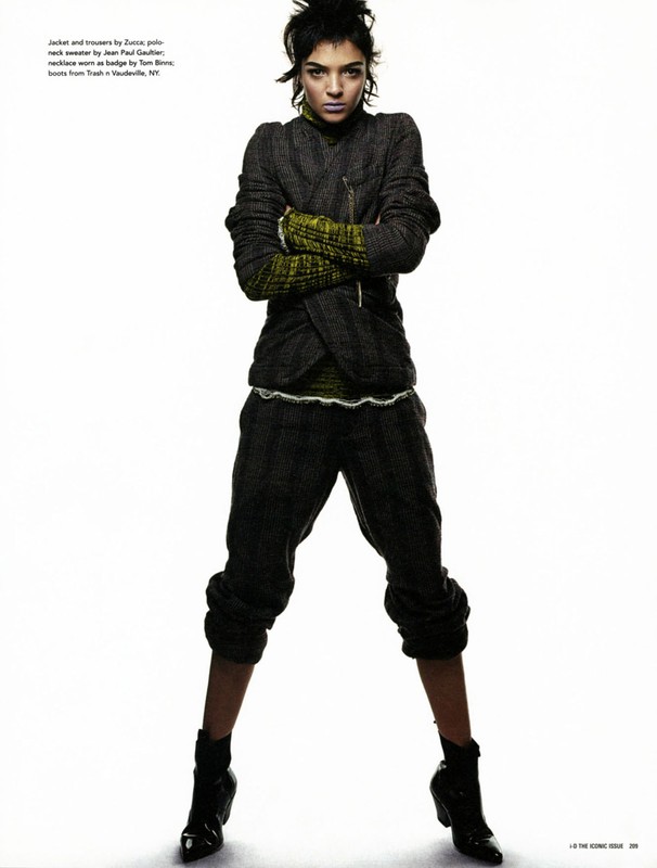 Fashion model in a gray jacket, green top, and gray pants with black boots standing against a white backdrop in a studio.