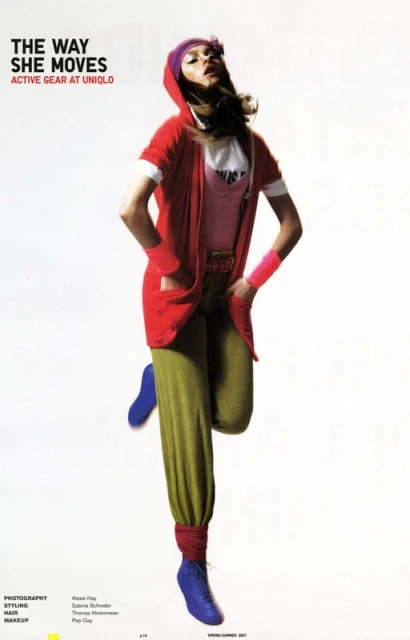 A woman in athletic wear stands on one leg in a studio. She is wearing red, pink, green, and blue clothing. She has on a head covering and wrist bands.
