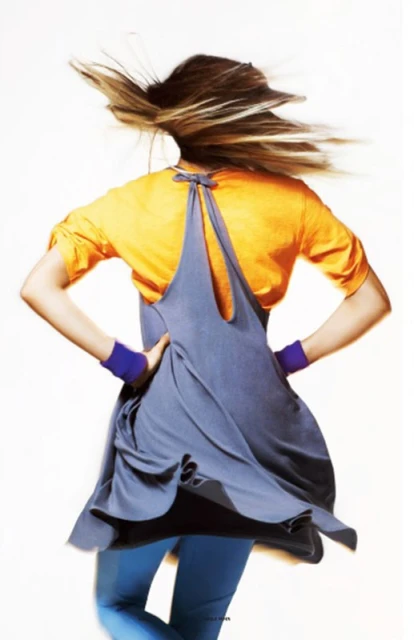 Woman in motion wears gray dress over orange shirt, purple wristbands, and blue leggings, captured against a bright white background.