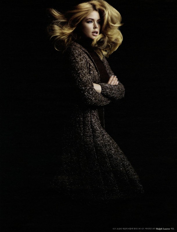 Fashion shot of a woman with blonde hair wearing a brown knit coat against a dark background in a studio, arms crossed.