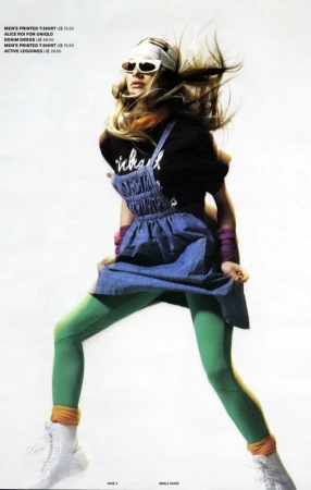 Fashion editorial shot of a woman in 80s style denim dress, green leggings, and white boots, striking a pose with windblown blonde hair.