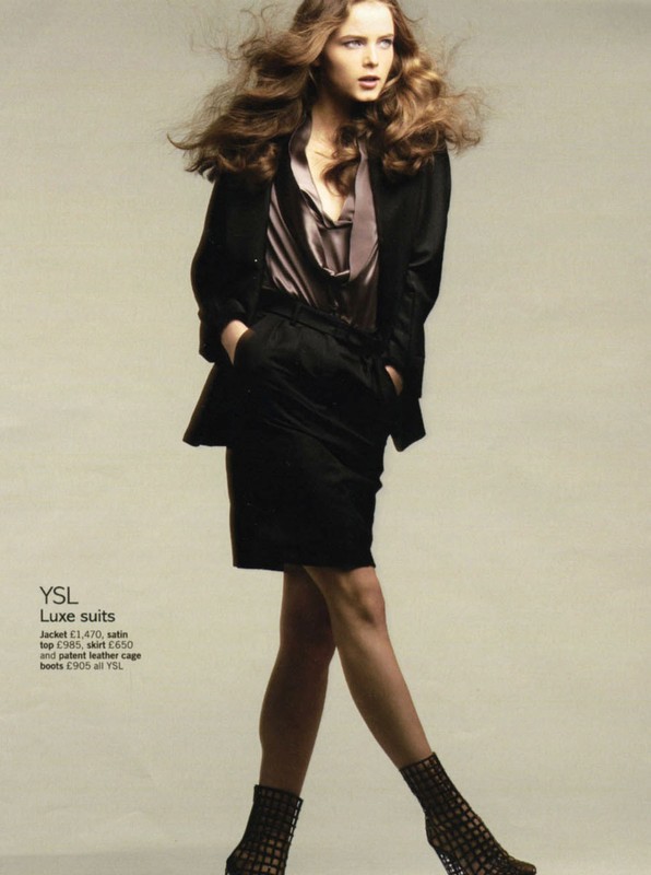 Fashion editorial featuring a woman with voluminous brown hair wearing a black skirt suit and geometric heels against a light grey backdrop.