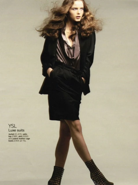 Fashion editorial featuring a woman with voluminous brown hair wearing a black skirt suit and geometric heels against a light grey backdrop.