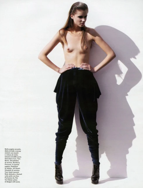 A nude fair-skinned model poses in a high-fashion, full-length studio shot wearing black harem pants and patent platform ankle boots, next to a shadow.
