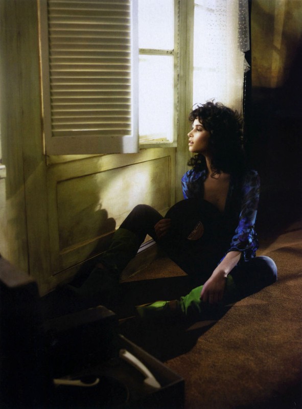 Adult woman sitting near a window in a dim room, holding a hat and wearing a floral-patterned shirt and boots. Indoor setting.