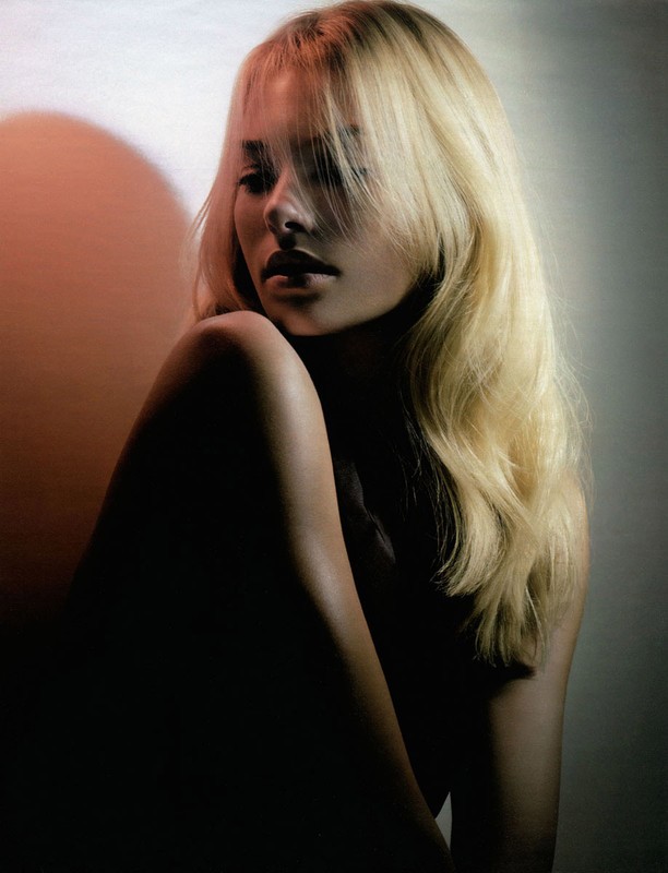 Portrait of a blonde woman with styled hair looking down in soft lighting with a warm circular light projection in background.
