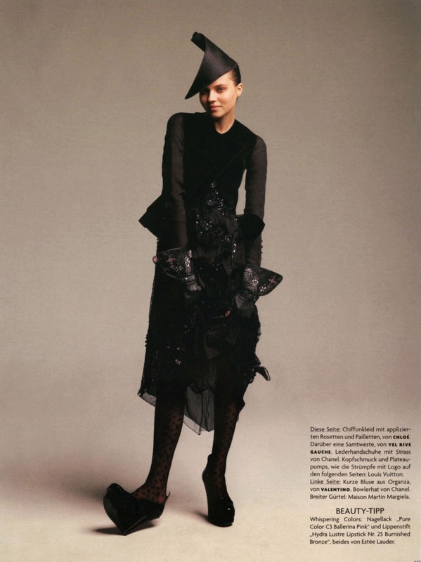 Model in black avant-garde dress wearing an asymmetrical hat and platform heels against a neutral backdrop