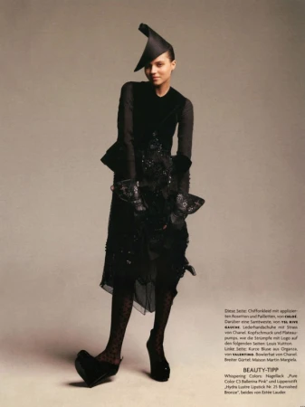 Model in black avant-garde dress wearing an asymmetrical hat and platform heels against a neutral backdrop