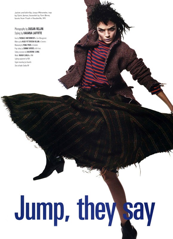 Fashion editorial shot of a woman jumping and wearing a striped shirt, brown jacket, black skirt, and ankle boots against white background.