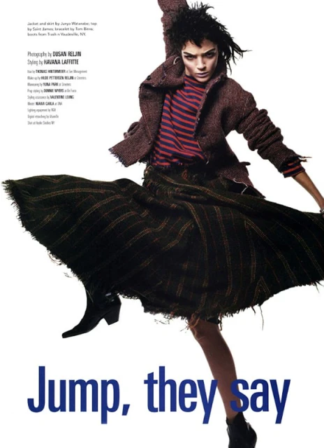 Fashion editorial shot of a woman jumping and wearing a striped shirt, brown jacket, black skirt, and ankle boots against white background.