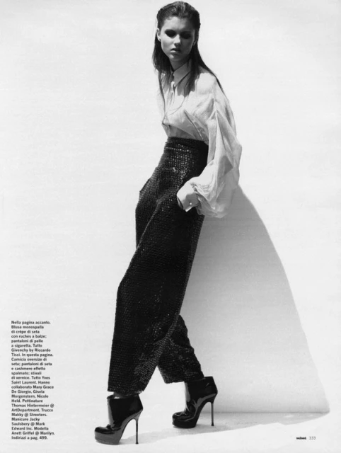 Fashion editorial shot of a young adult woman with dark hair wearing high-waisted sequin pants, a blouse, and platform heels in a studio.