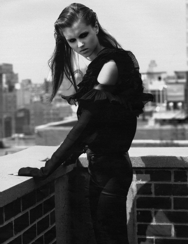 Model with slicked-back hair in a black ruffled top leaning on a brick rooftop ledge against an urban skyline background in black and white.