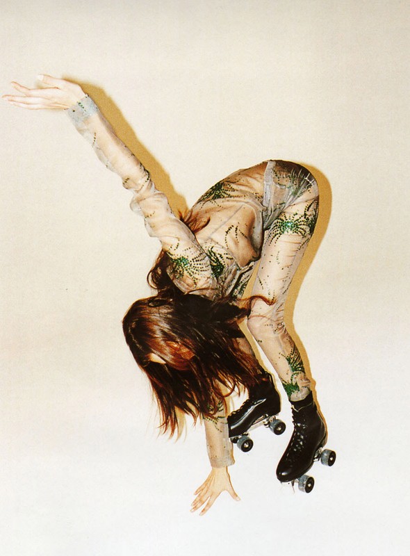 Woman in patterned mesh catsuit bends forward on roller skates in a studio, reaching out with one arm against a plain cream background.