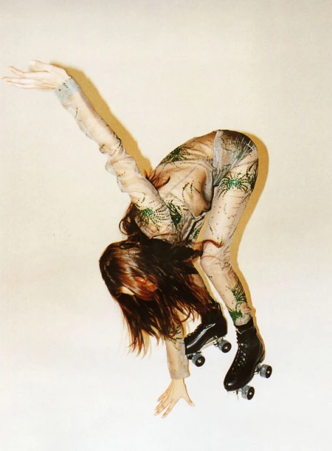 Woman in patterned mesh catsuit bends forward on roller skates in a studio, reaching out with one arm against a plain cream background.