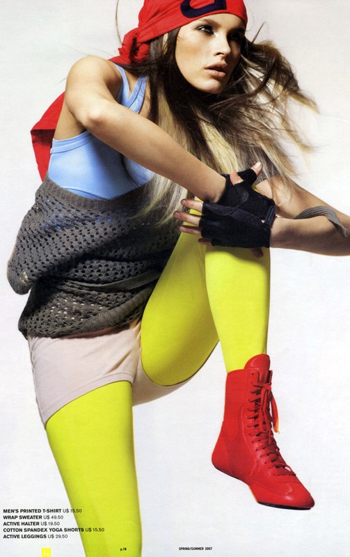 Athletic woman in red cap and yellow leggings stretches her leg, wearing red boots and blue top against a plain studio background.
