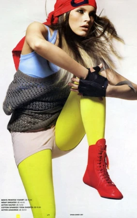 Athletic woman in red cap and yellow leggings stretches her leg, wearing red boots and blue top against a plain studio background.