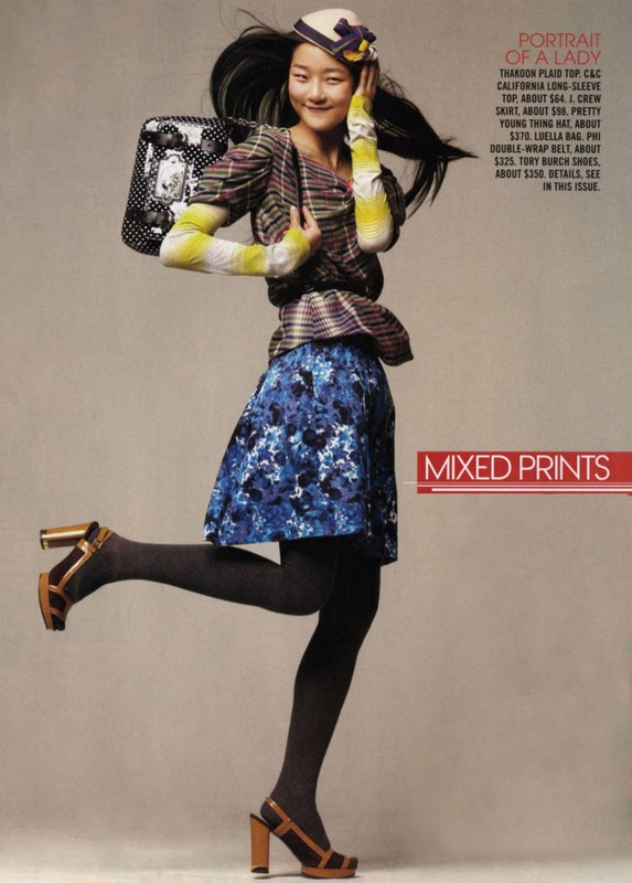 Asian female model strikes a pose in a stylish, mixed-print ensemble featuring a floral skirt, plaid top, and unique handbag, against a neutral backdrop.