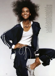 Smiling Black woman with voluminous curly hair wearing a dark navy tracksuit and white tank top against a plain grey background