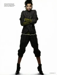 Model standing with arms crossed wearing a dark textured jacket and trousers against a white seamless backdrop.